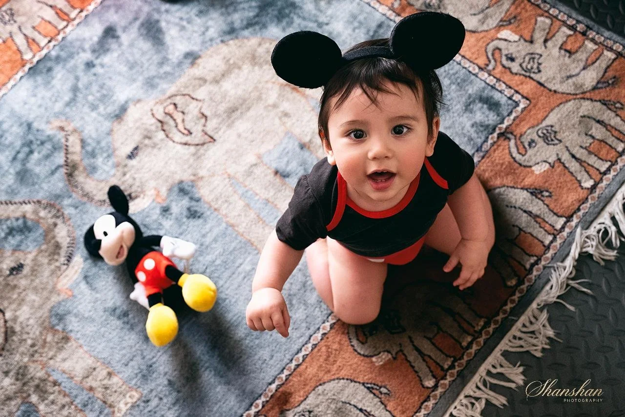 A toddler wearing Mickey Mouse ears and a black and red outfit, sitting on a patterned rug, looking up at the camera. A Mickey Mouse plush toy lies on the rug nearby.