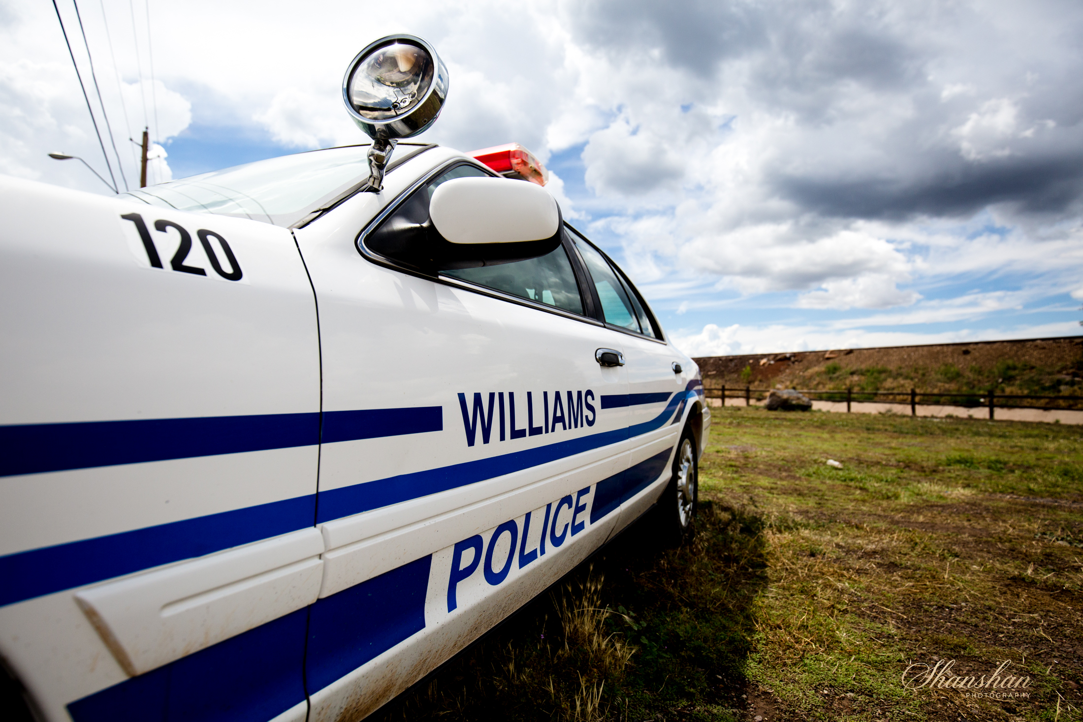 A white police car with blue stripes and the word WILLIAMS on the side, parked on grass under cloudy skies.