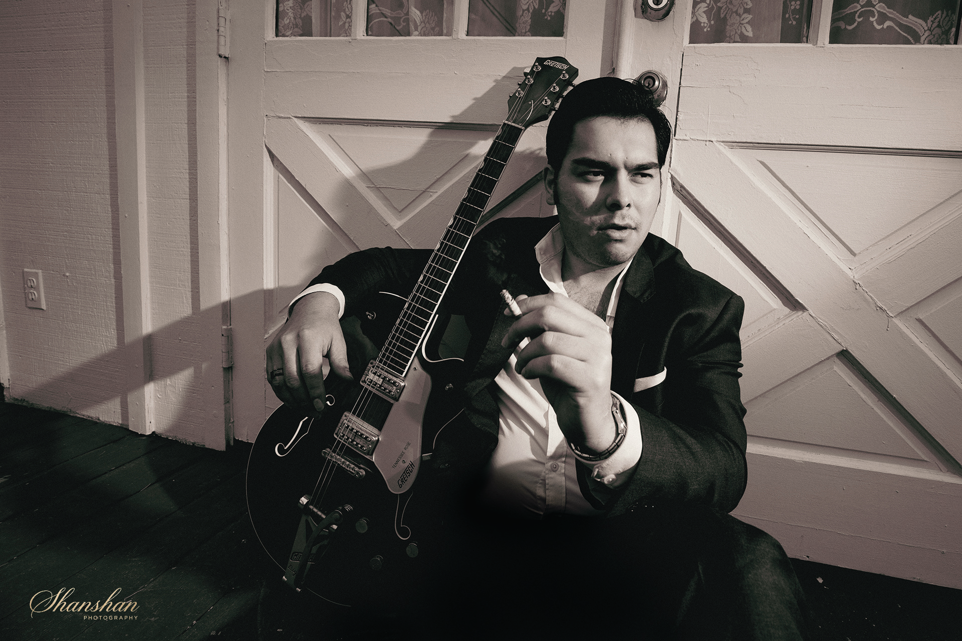 A black and white photo of a man sitting on a wooden floor against a white paneled door, holding an electric guitar and smoking a cigarette, with a contemplative expression.