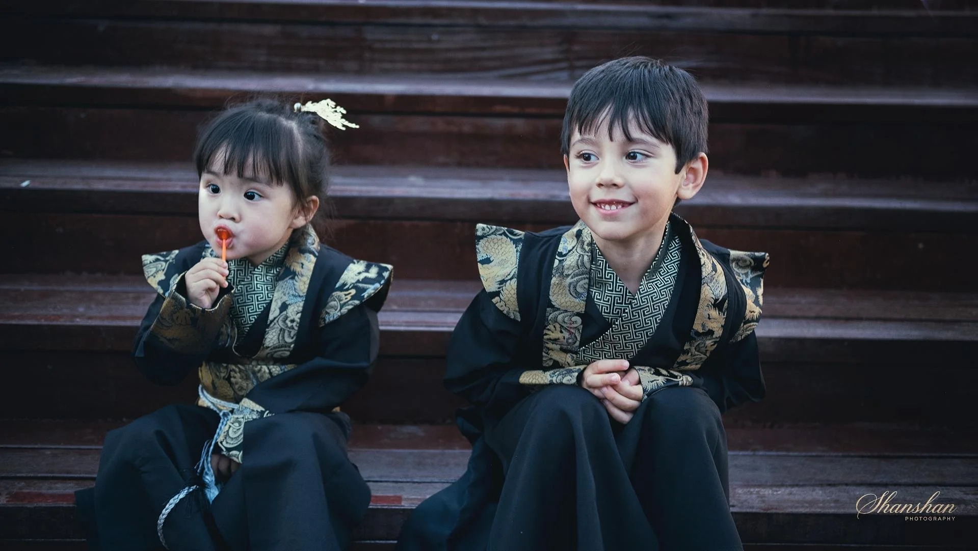 Two children sitting on wooden stairs, dressed in traditional black and gold attire, one girl licking a lollipop and a boy smiling with his hands clasped.