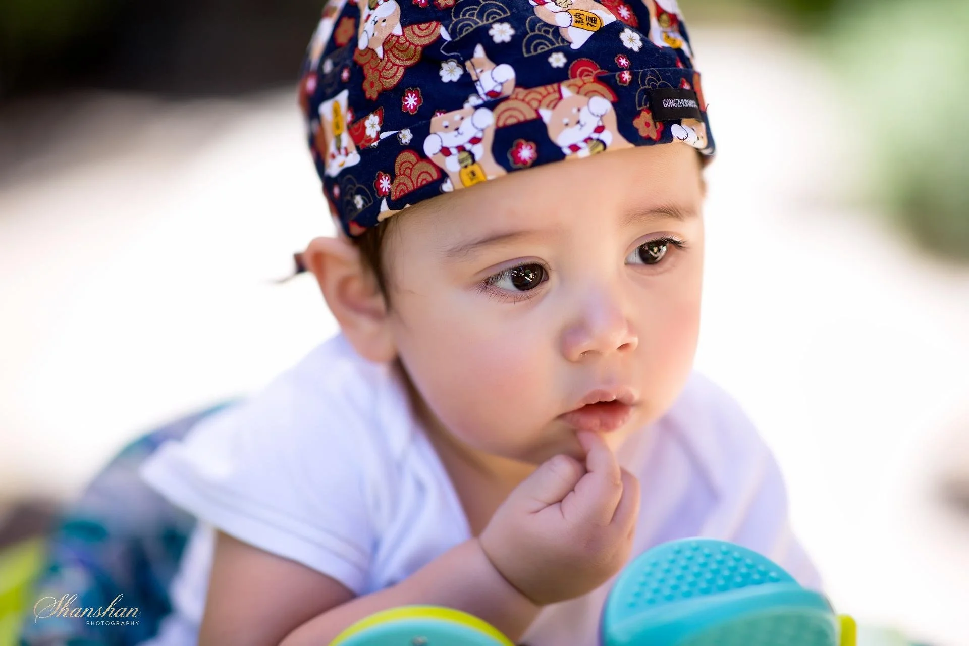 Close-up of a young child with light skin, dark eyes, and brown hair wearing a colorful hat with traditional Japanese patterns. The child is resting on their hands, appearing to be outdoors with a blurred background, and looking thoughtfully.