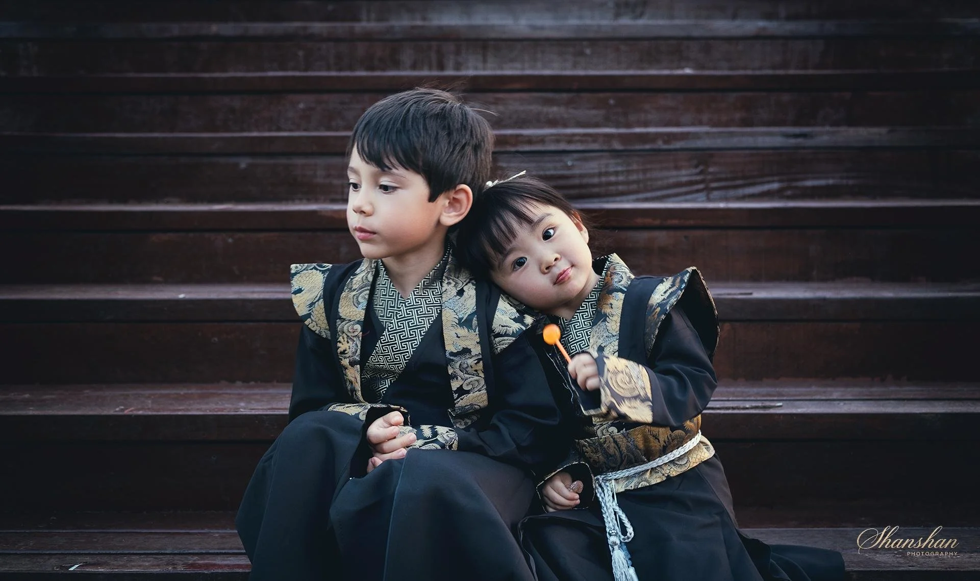 Two children dressed in traditional Japanese kimonos sitting on wooden stairs, one child resting head on the other's shoulder, holding a small orange and black toy.