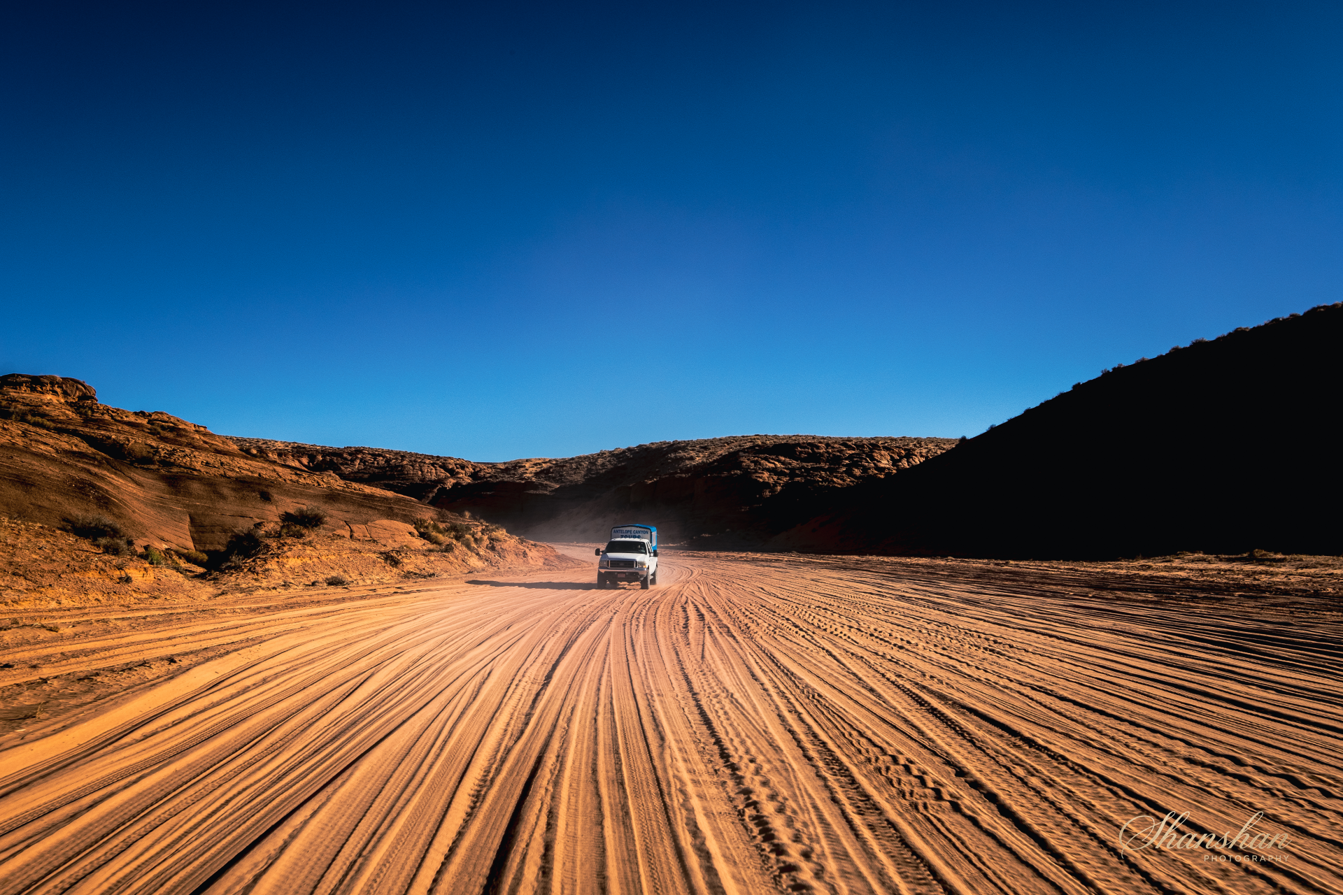 A white vehicle driving on a dirt road through a desert landscape with rocky hills on either side under a clear blue sky.