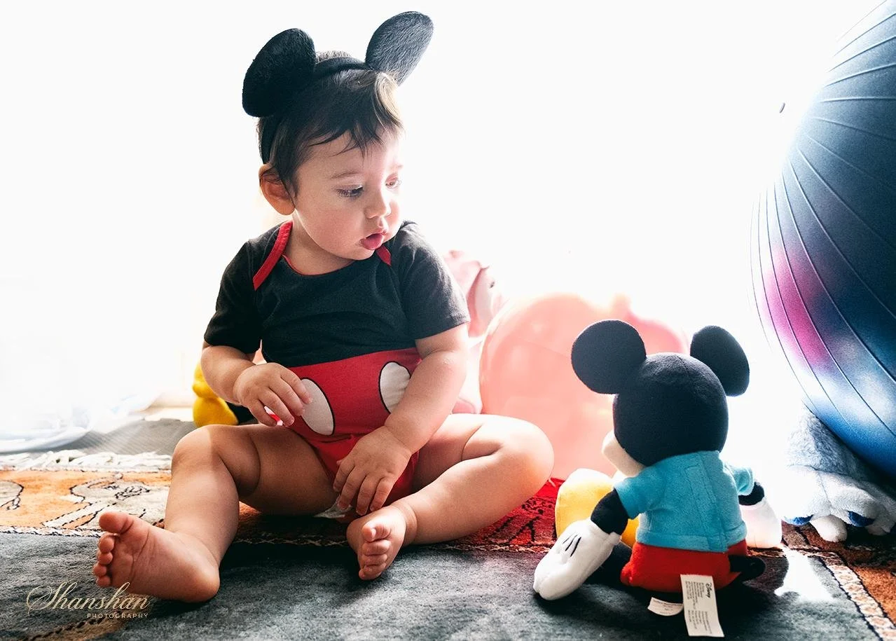 A young child wearing a Mickey Mouse costume and mouse ears hat, sitting on a carpet, looking at a Mickey Mouse plush toy, surrounded by balloons.