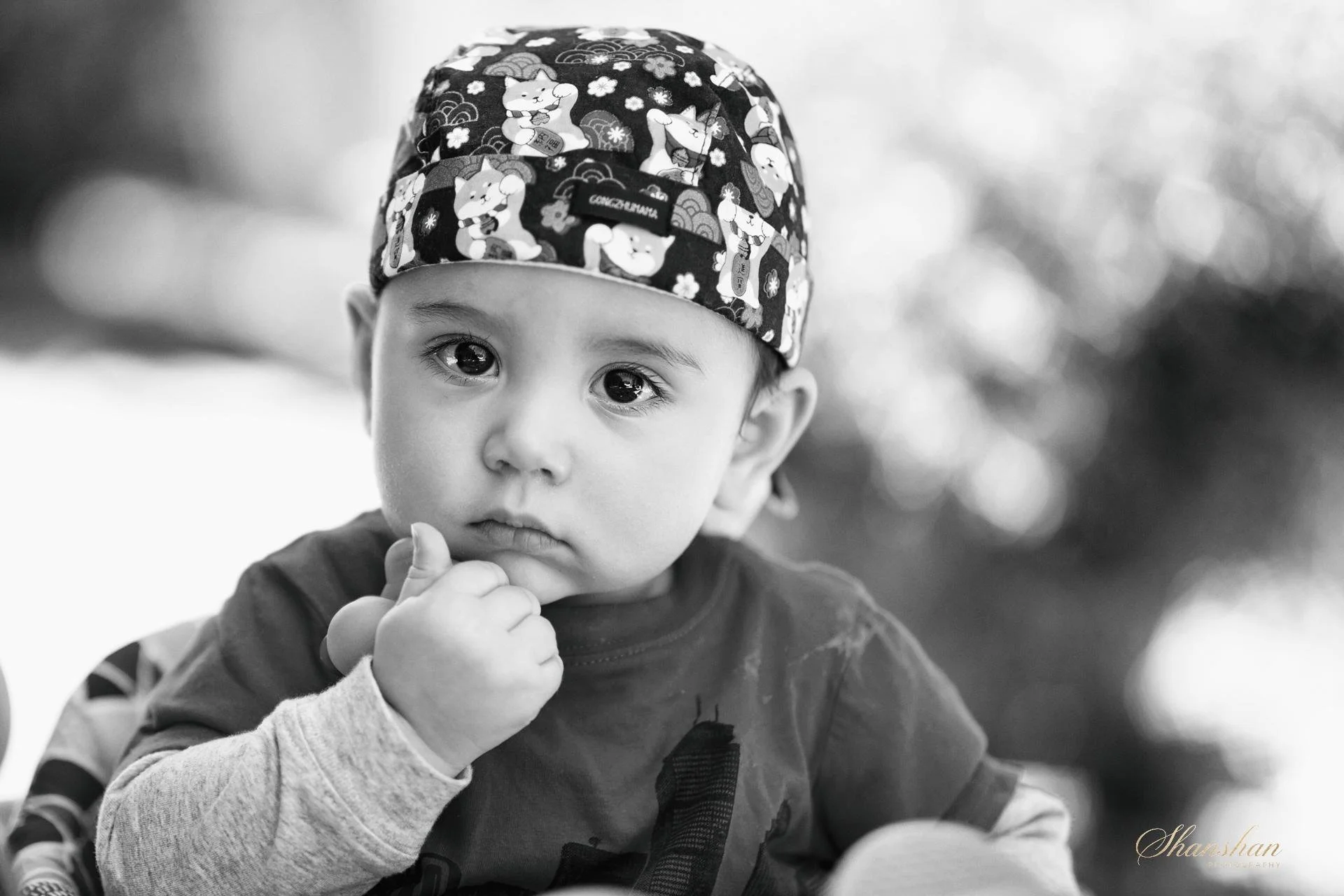 A young child with big eyes wearing a patterned cap and a long-sleeved shirt, holding his hand near his mouth and looking off to the side.