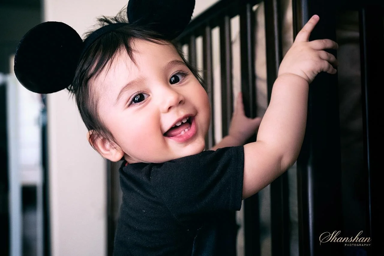 A smiling young girl with black hair wearing black Mickey Mouse ears, holding onto a black railing.