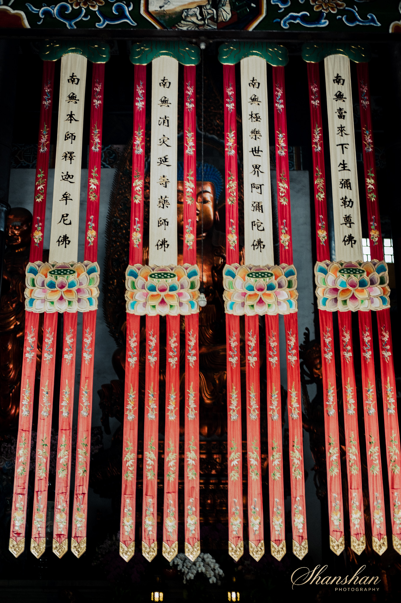 Decorative hanging prayer flags with Chinese characters and colorful floral patterns, with a Buddha statue in the background.