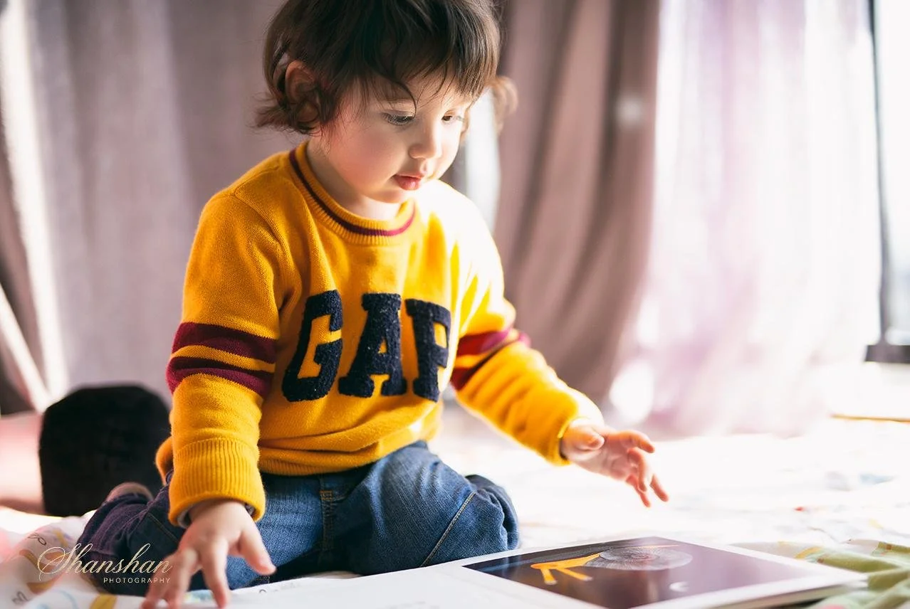 Young child sitting on bed, wearing a yellow GAP sweater, looking at photos or a book on the bed.