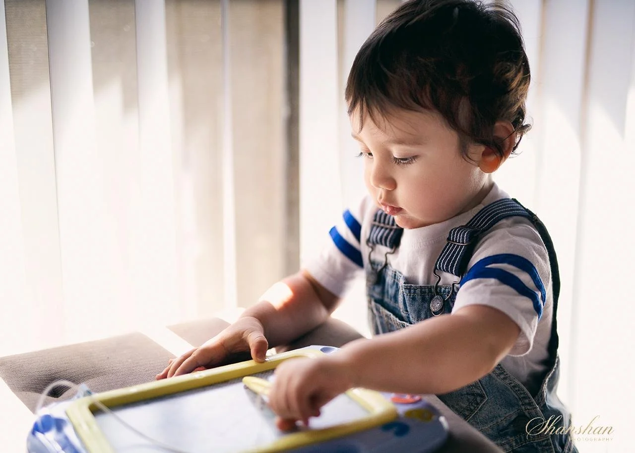 A young boy with dark hair, wearing a white shirt with blue stripes and denim overalls, playing with a yellow magnetic drawing board at a table near a bright window.