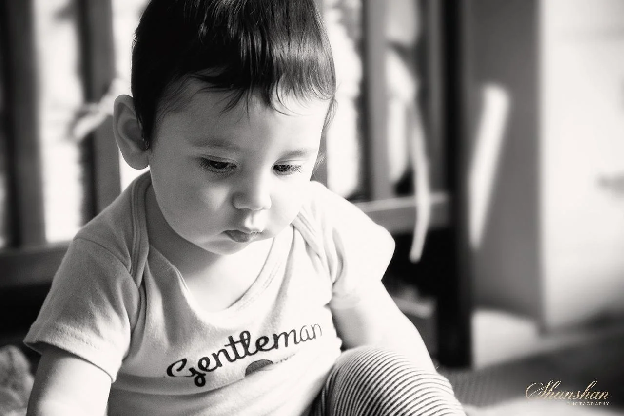 A young boy with short dark hair looking down with a serious expression, sitting indoors.