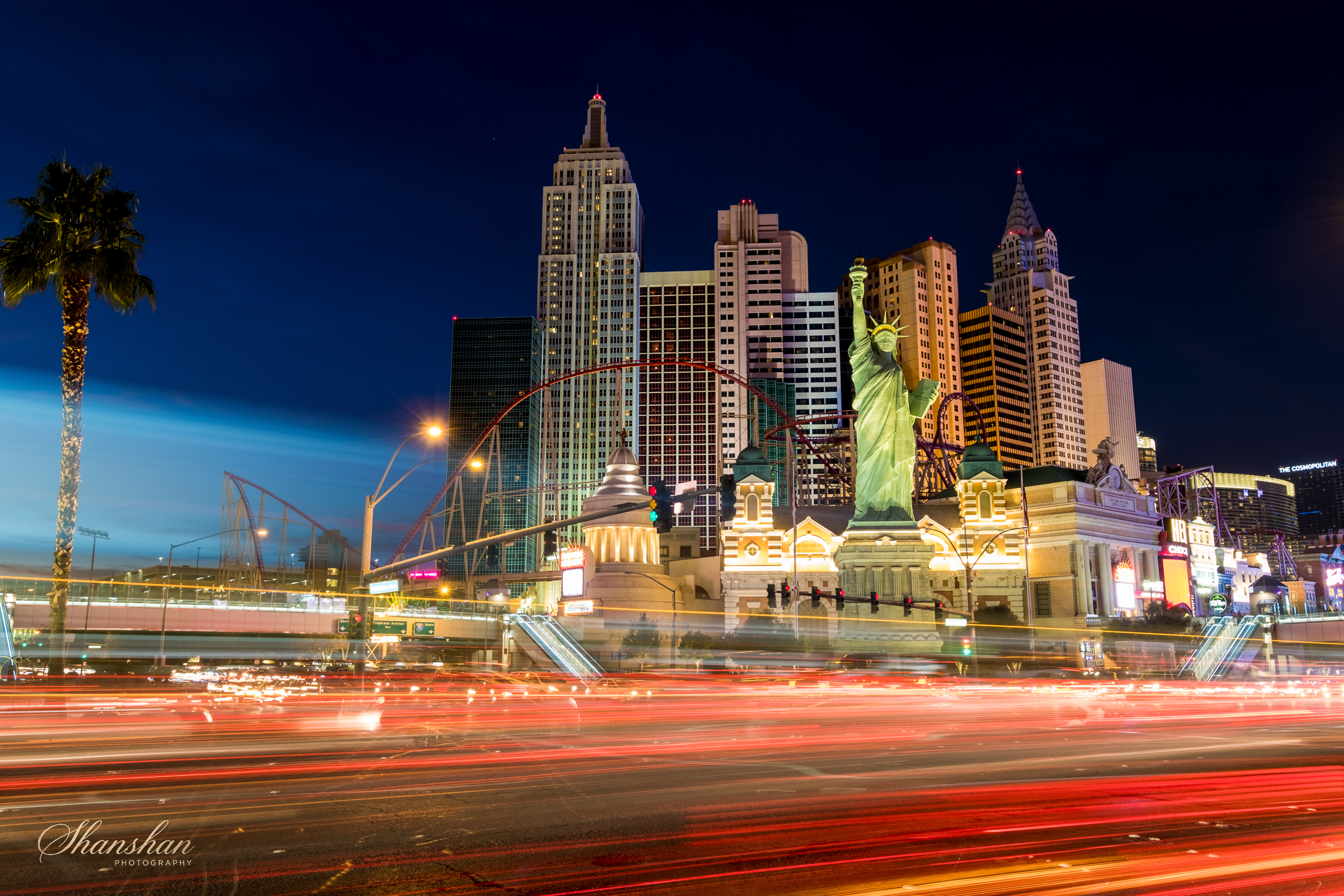 Nighttime scene of the Las Vegas Strip featuring the New York-New York Hotel and Casino with replica of the Statue of Liberty, illuminated tall skyscrapers, a roller coaster, and light trails from moving vehicles.