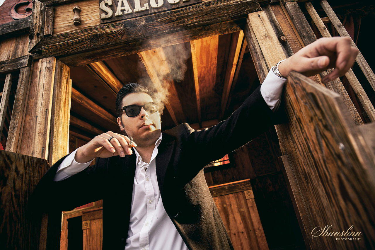 A man in black sunglasses, a black blazer, and a white shirt smokes a cigarette inside a wooden saloon structure.