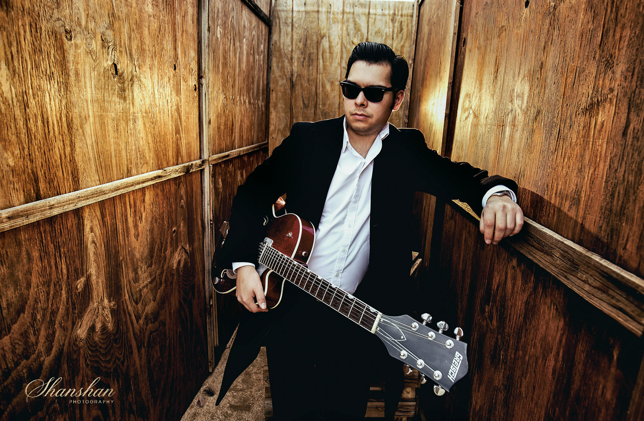 A man dressed in a black suit and white shirt, wearing sunglasses, leaning against wooden panel walls while holding an electric guitar.