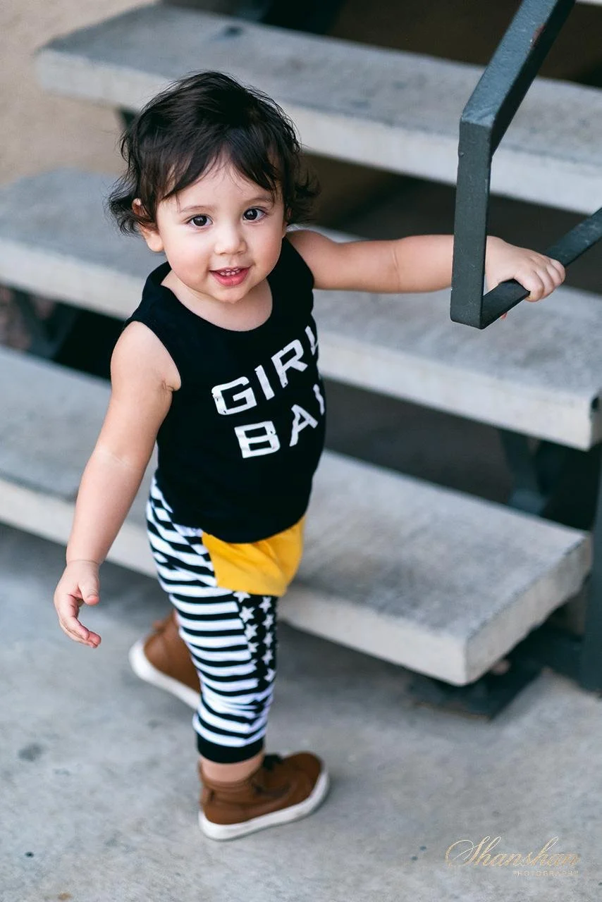 A young girl with dark hair, wearing a black sleeveless shirt that says 'GIRL BAD', striped black and white pants, and brown shoes, holding onto a metal railing on outdoor stairs and looking up at the camera with a smile.