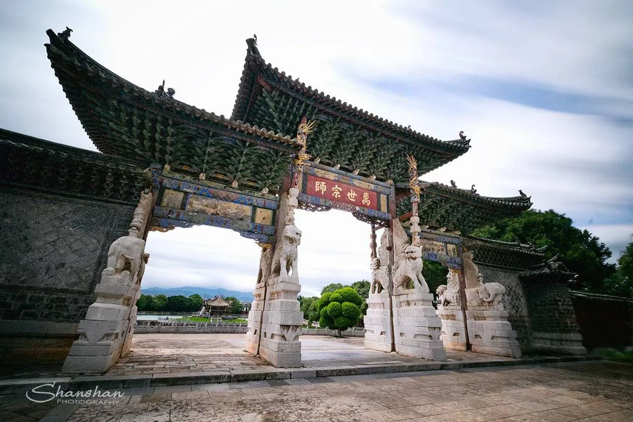 Traditional Chinese-style gate with ornate carvings, lion statues, and a tiled roof, set in a park with trees and a lake in the background.