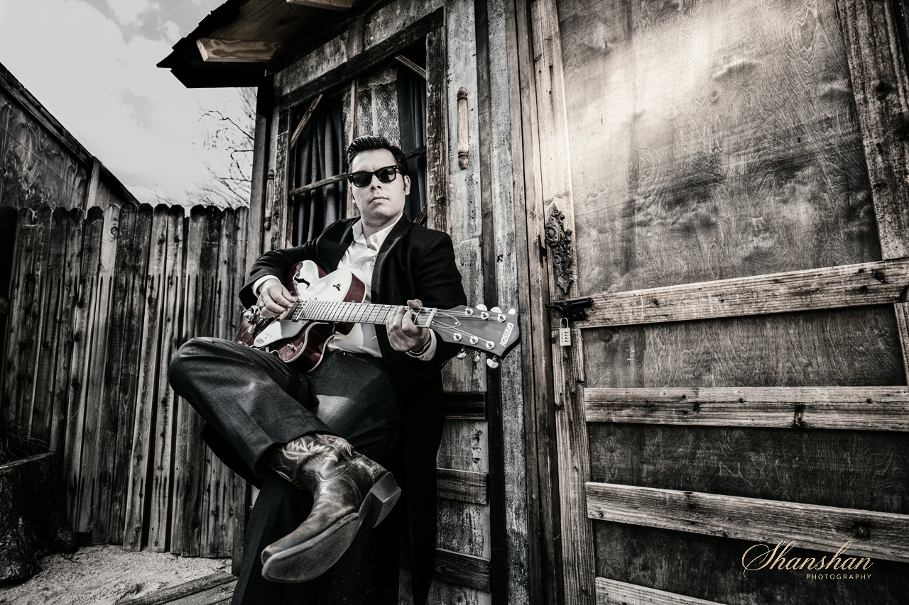 A man wearing sunglasses, a black coat, and cowboy boots sitting against a wooden fence, playing an acoustic guitar outside near a wooden building.