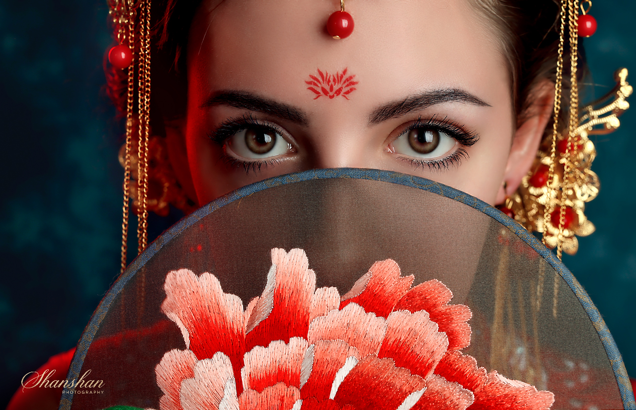 A woman with traditional Asian jewelry and makeup, partially hiding her face behind a fabric fan with floral embroidery, focusing on her brown eyes and forehead decoration.