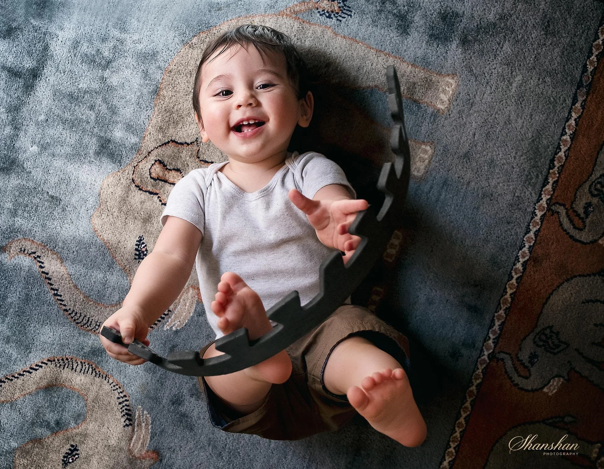 A smiling young boy lying on a colorful patterned carpet, holding a black frame or object, with a playful expression and missing front teeth.