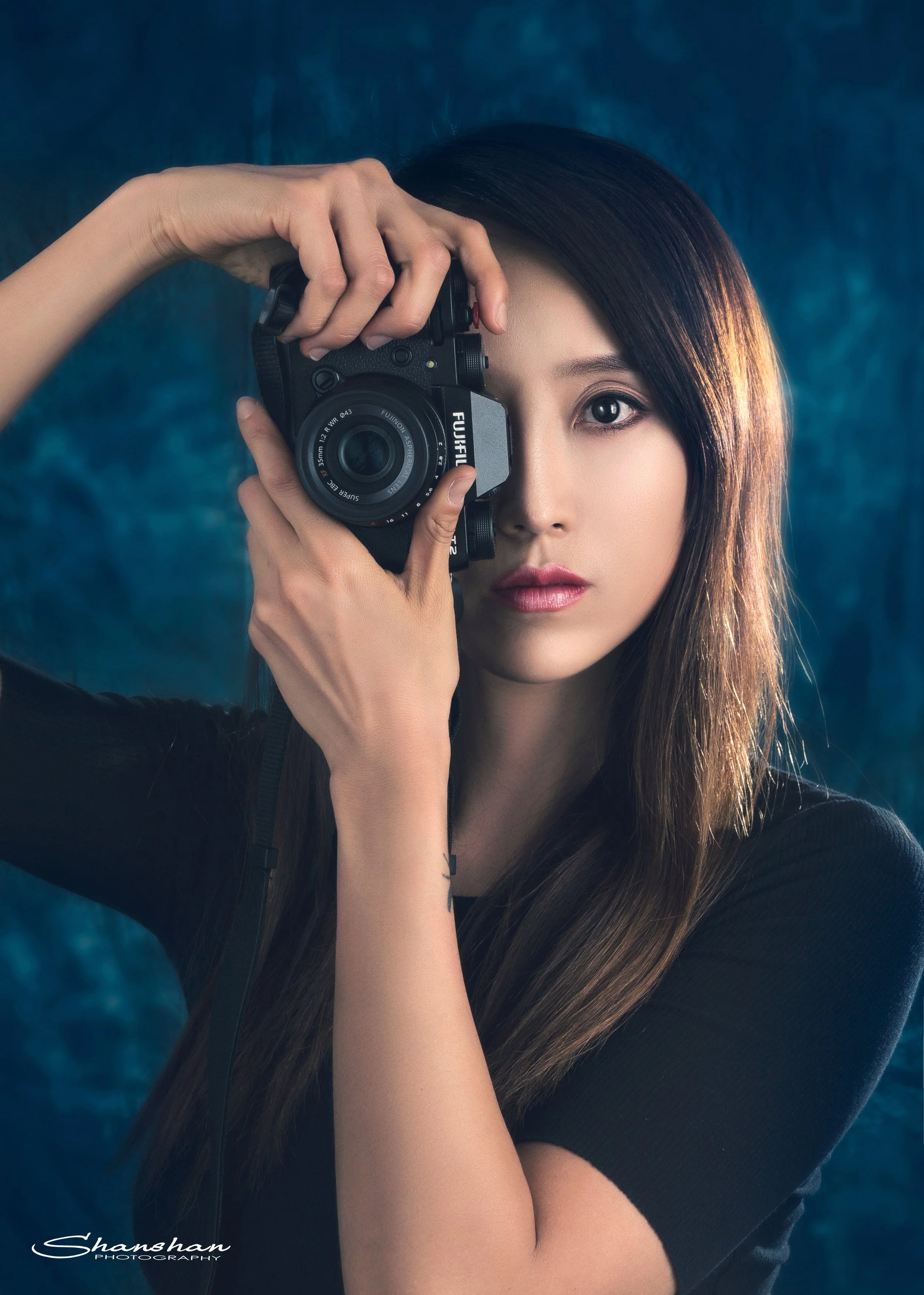 A woman with straight dark hair and light skin, holding a Fujifilm camera up to her eye, looking directly at the camera, against a dark blue textured background.