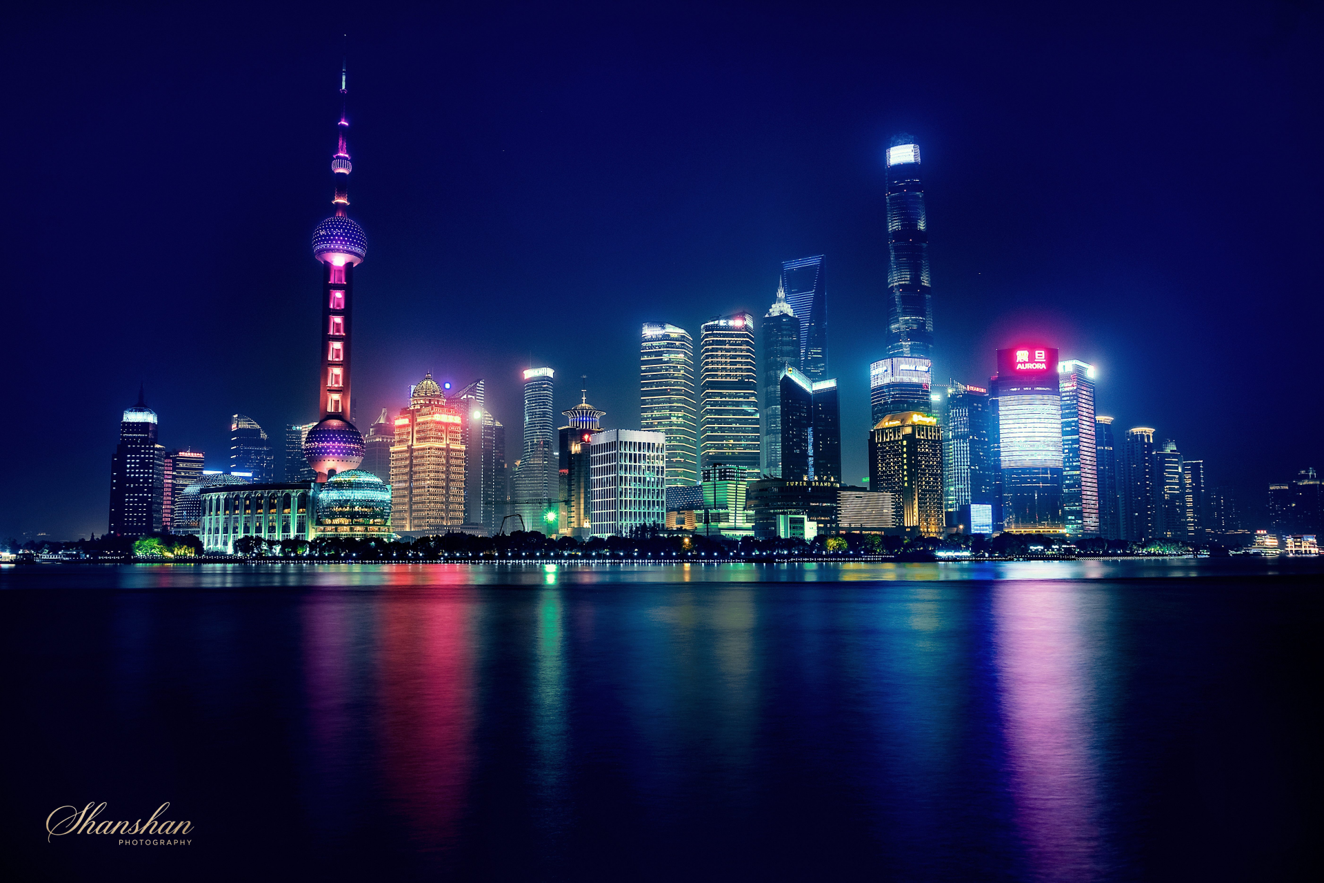 Night view of a city skyline with illuminated skyscrapers and the Oriental Pearl Tower, reflected on a river in Shanghai, China.