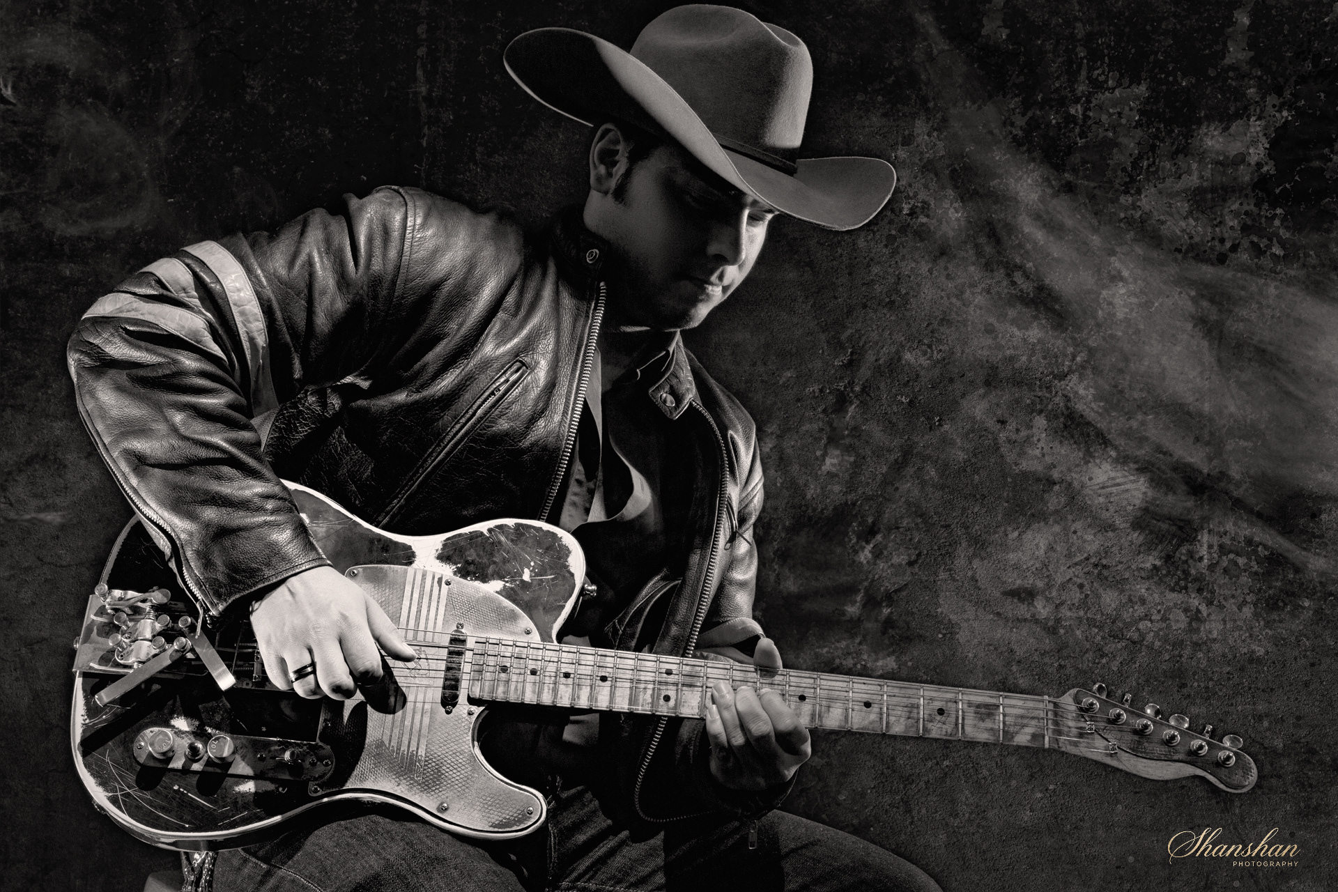 A black and white photo of a man wearing a hat and leather jacket playing an electric guitar with a textured wall background.
