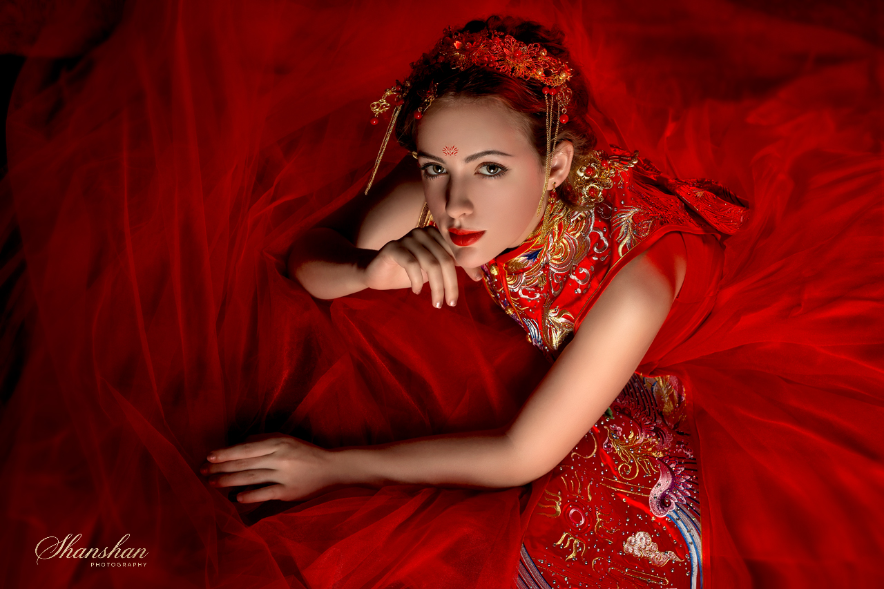 A woman dressed in a traditional red Chinese wedding gown with gold embroidery, lying on a red fabric background, with her head resting on her hand and looking at the camera.