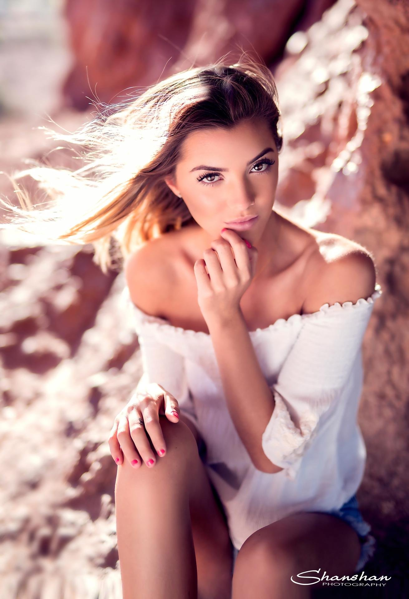 A young woman with long hair looking into the camera, wearing an off-shoulder white top, sitting on sandy ground near rocks with sunlight highlighting her hair.