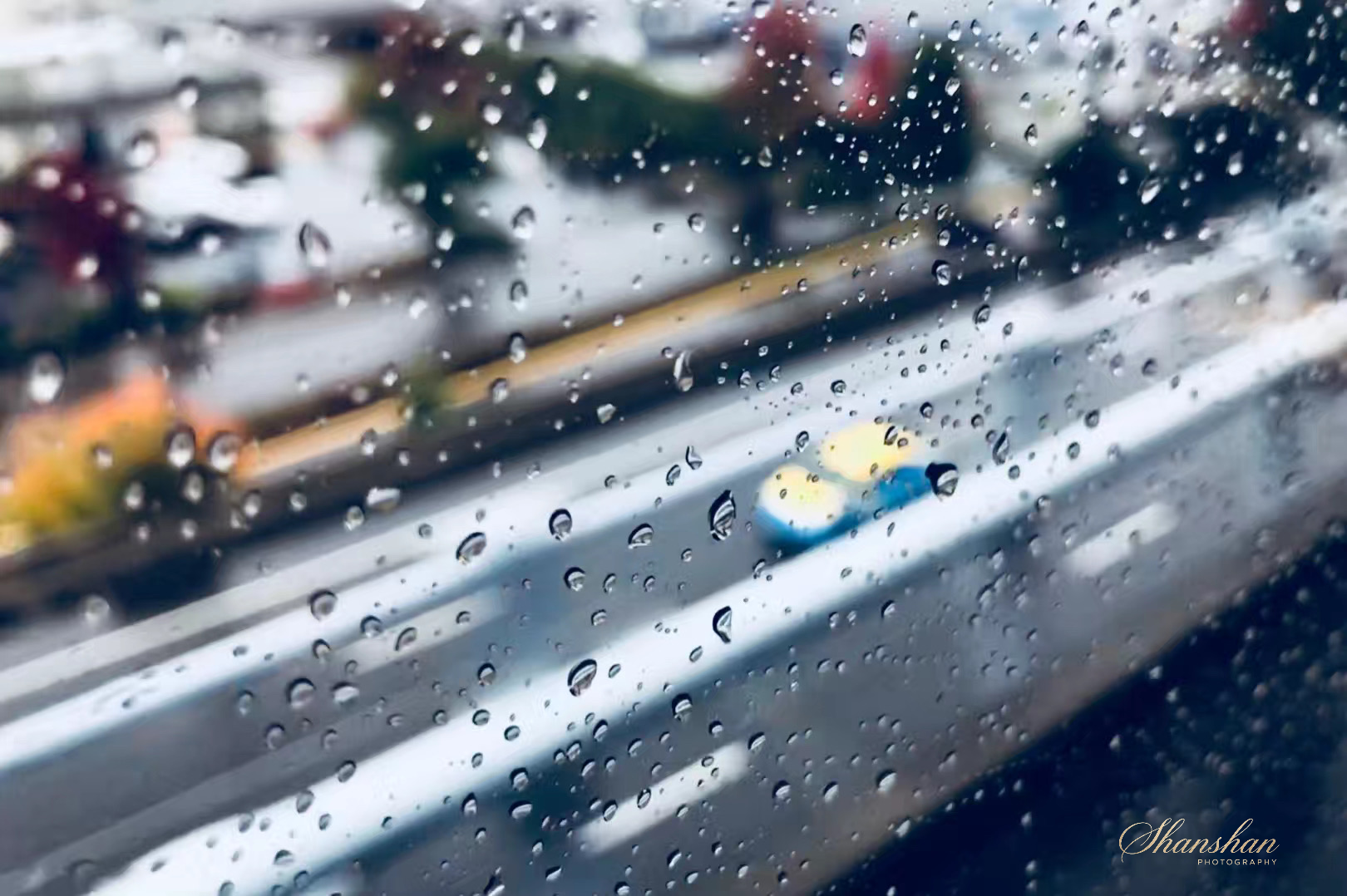 View of a rainy city street through a window with raindrops on it, showing blurred cars and buildings in the background.