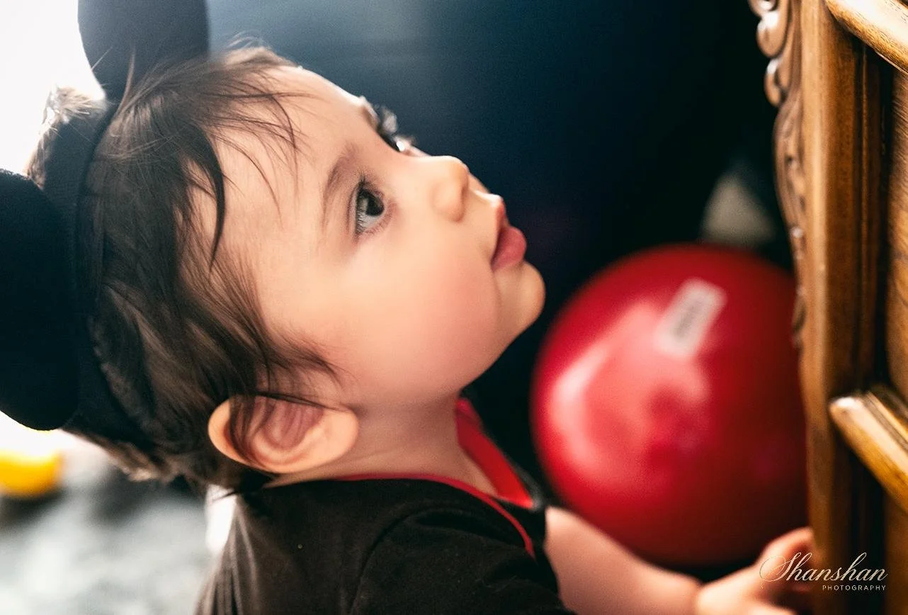 A young girl with dark hair, wearing a black headband and black shirt, looks up at a red ball in front of a wooden drawer.
