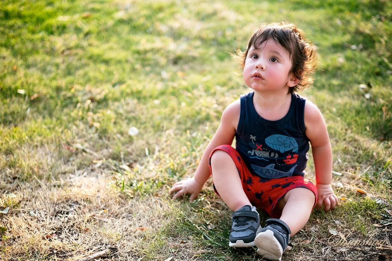 Young child sitting on the grass outdoors, wearing a sleeveless navy shirt, red shorts, and sneakers, with short messy hair and a curious expression.