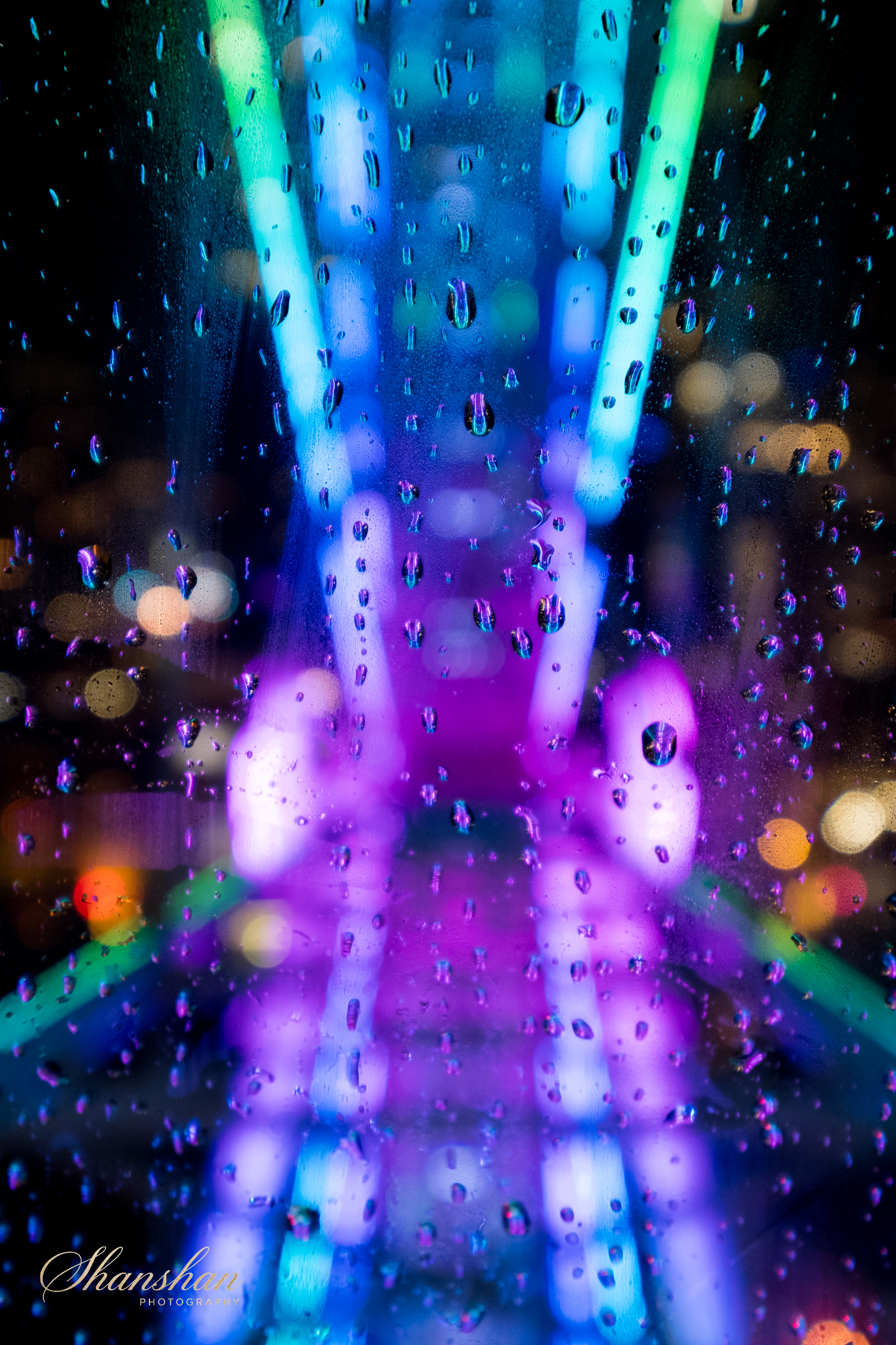 Colorful neon lights blurred behind a wet glass with raindrops, creating a vibrant, abstract scene.