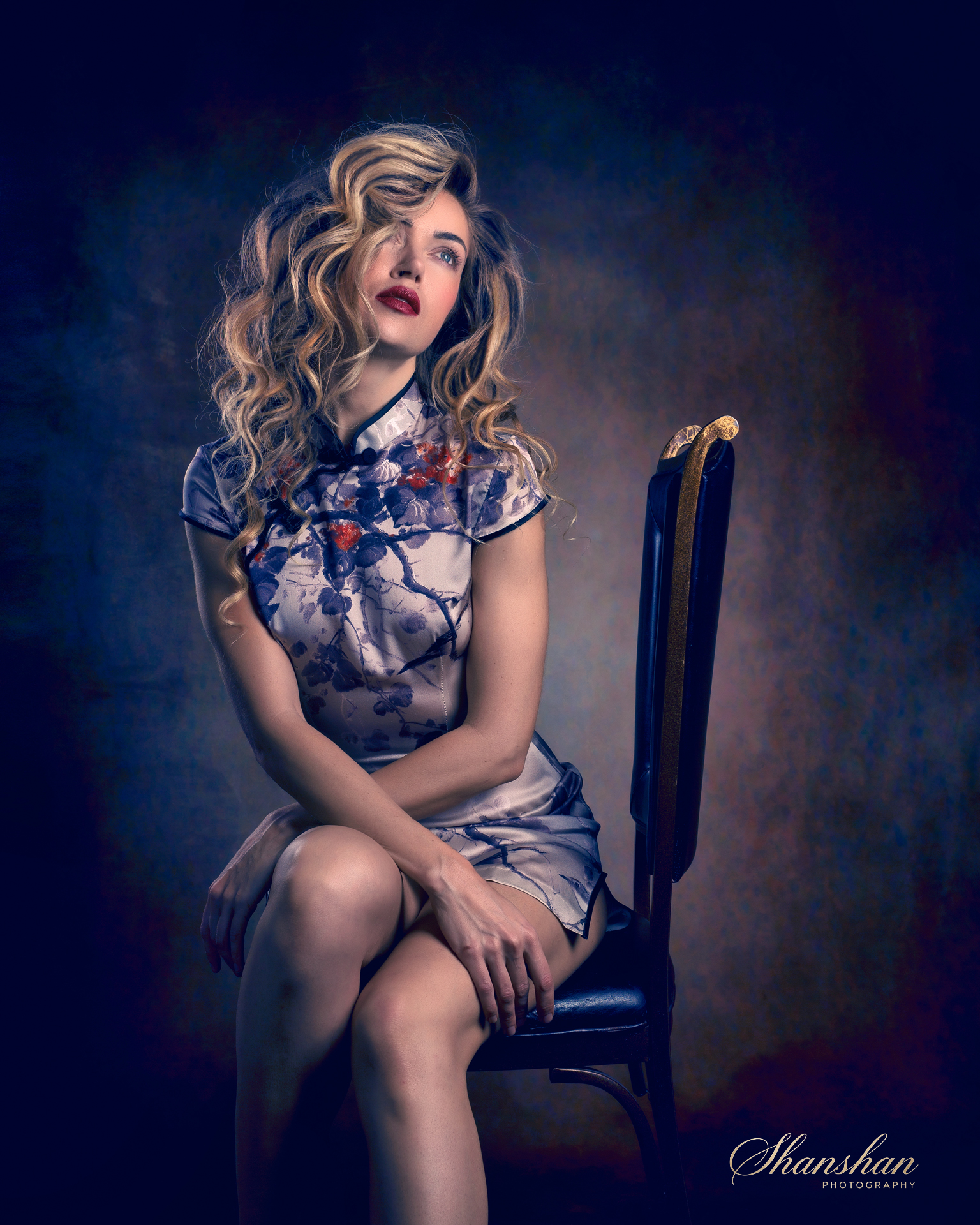 A woman with long curly blonde hair and dark red lipstick sitting on a black chair, wearing a floral dress, against a dark multicolored background.
