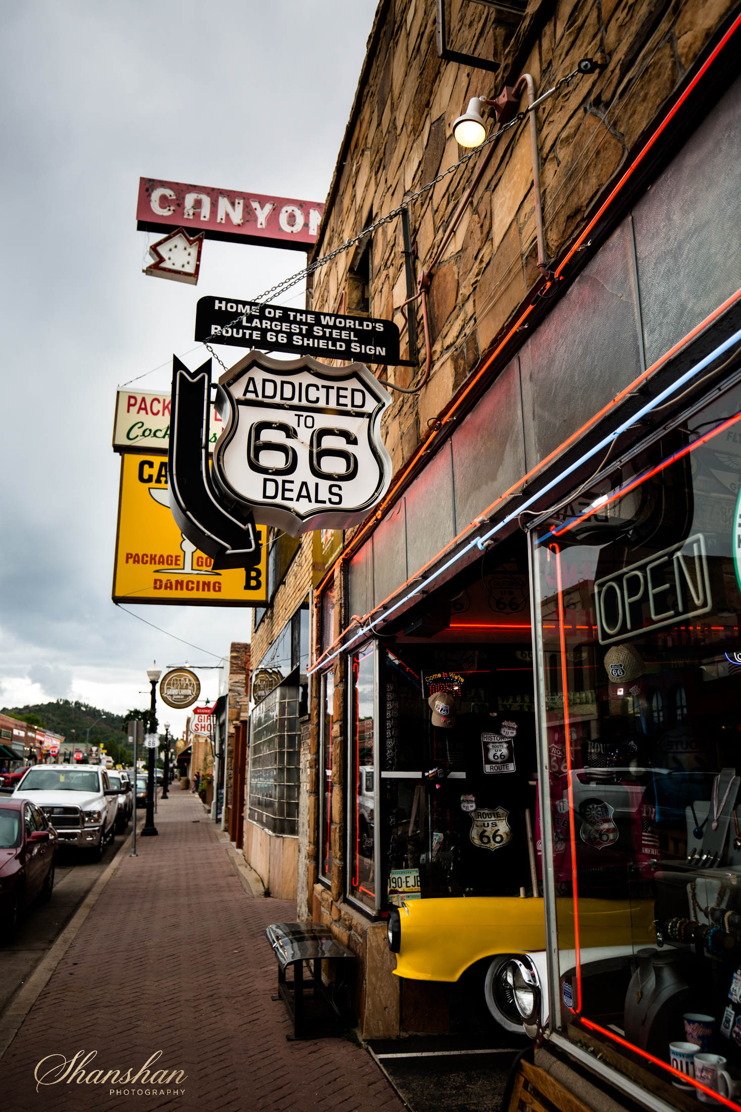 A street view featuring storefront signs including a neon Route 66 sign, a Canyon sign, and a sign indicating the home of the world's largest steel Route 66 shield sign. The storefront has a display window with various Route 66 memorabilia and neon l