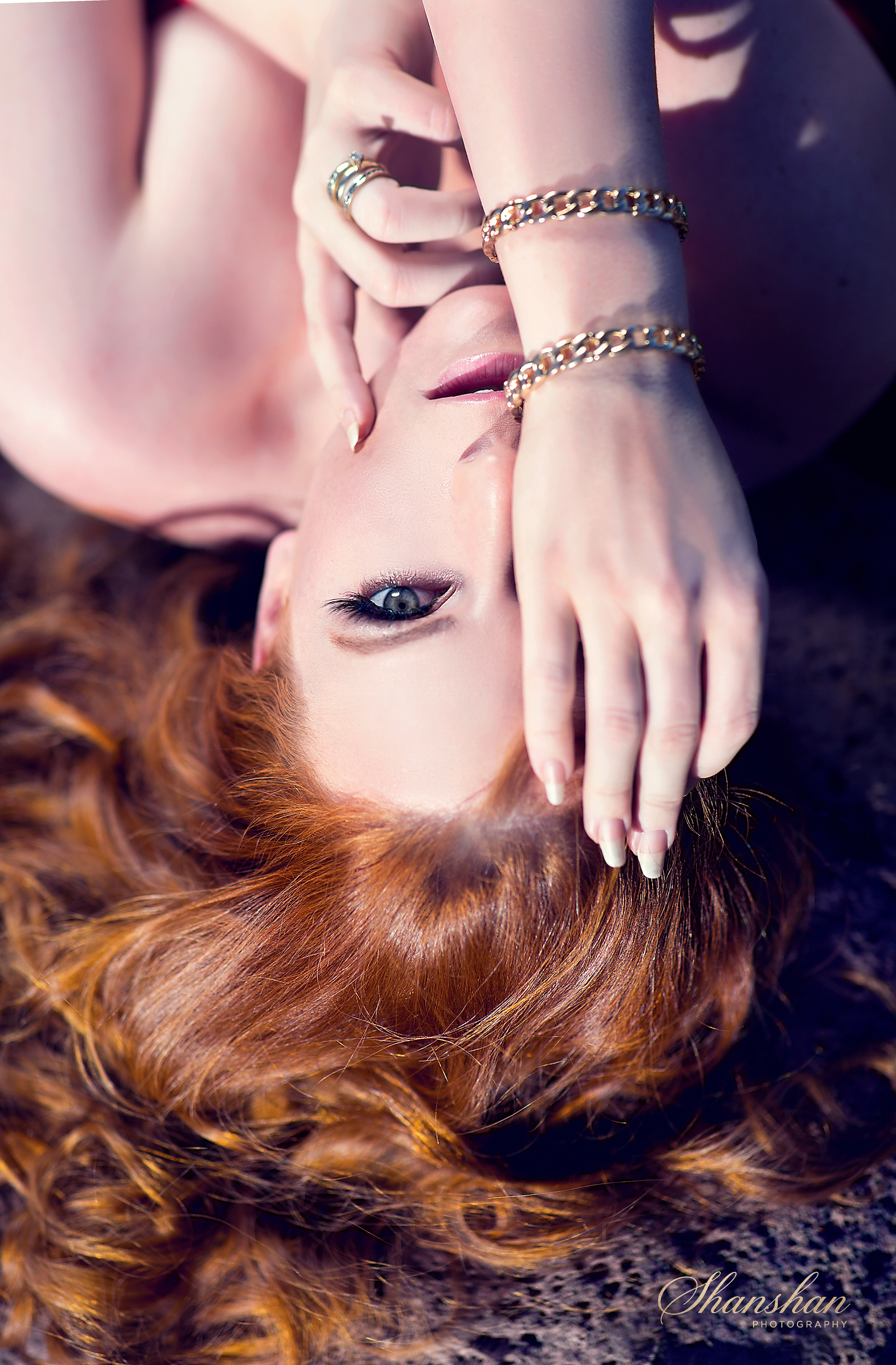 A woman with red curly hair lying on the ground, covering part of her face with her hand, wearing rings and bracelets, with her eye visible and looking at the camera.