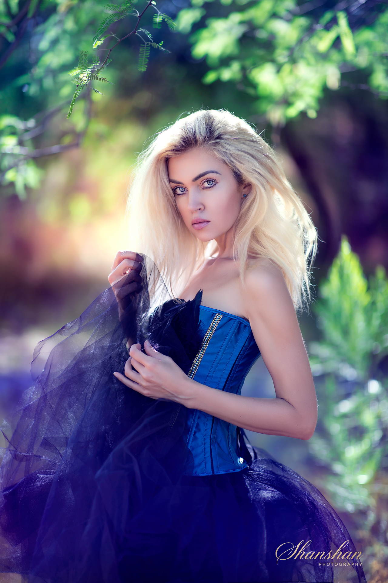 A woman with blonde hair and blue eyes wearing a blue dress with black tulle fabric, standing outdoors among green trees and foliage, gazing at the camera with a serene expression.
