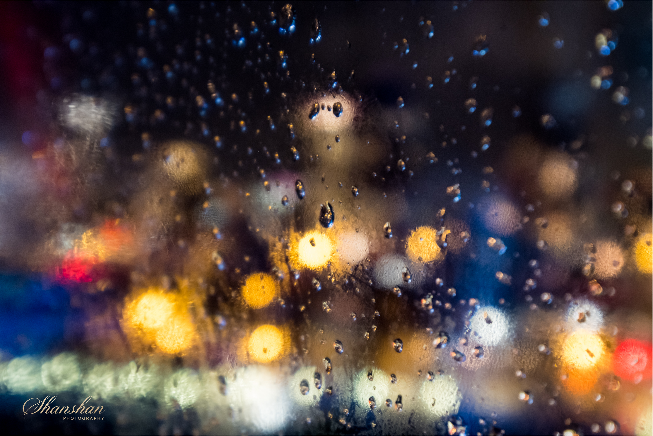 Close-up of a foggy or wet glass surface with numerous water droplets, with blurred city lights in the background creating bokeh effects in various colors, including yellow, blue, red, and white.