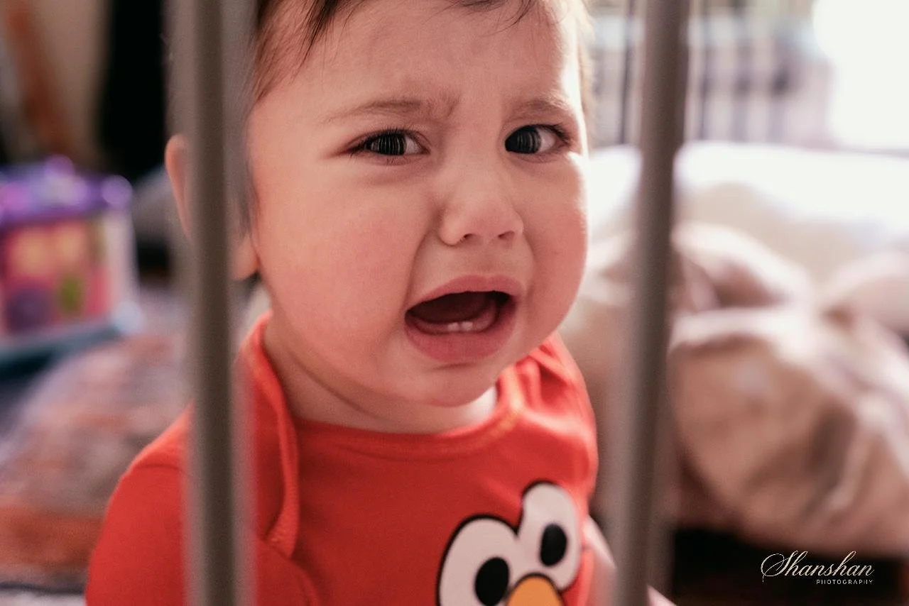 A young child with a distressed facial expression, crying behind bars, wearing a red shirt with a cartoon character on it.