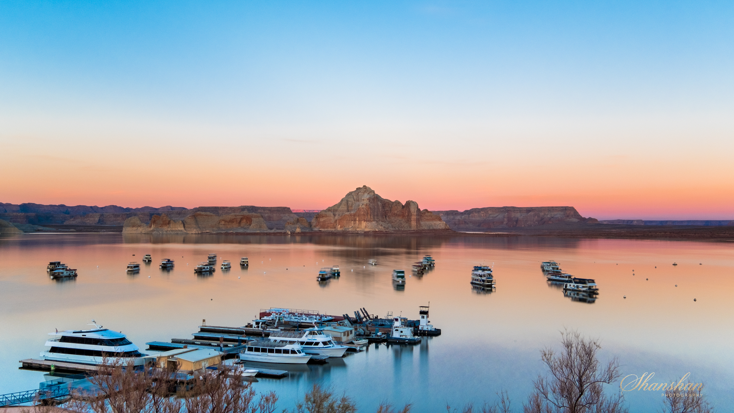 Calm lake reflecting colorful sunset sky, with boats anchored, and layered rock formations in the distance.