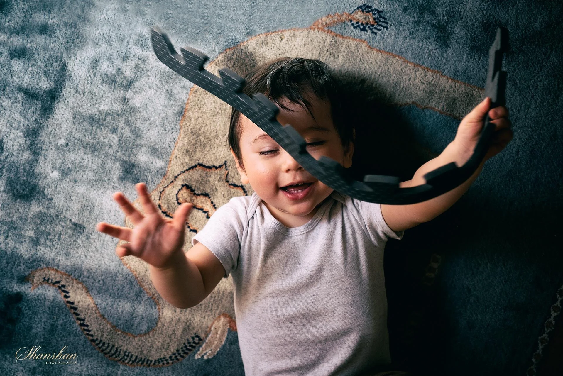 Young child lying on a colorful patterned rug, smiling and holding a black toy sword, with the sword's blade resting near their face.