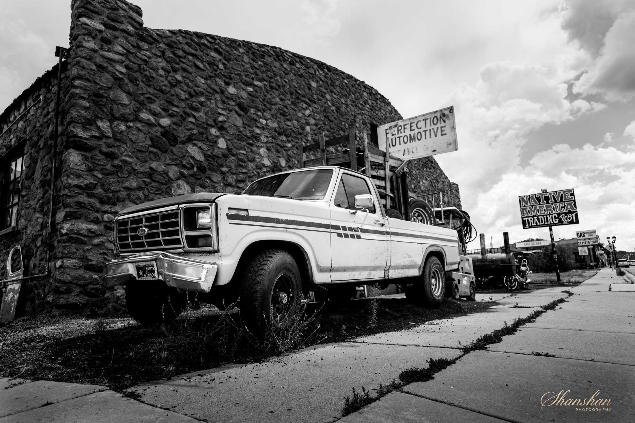 A black and white photo of a vintage Ford pickup truck parked on a sidewalk in front of a stone building. There are signs in the background advertising Native American trading and automotive services.