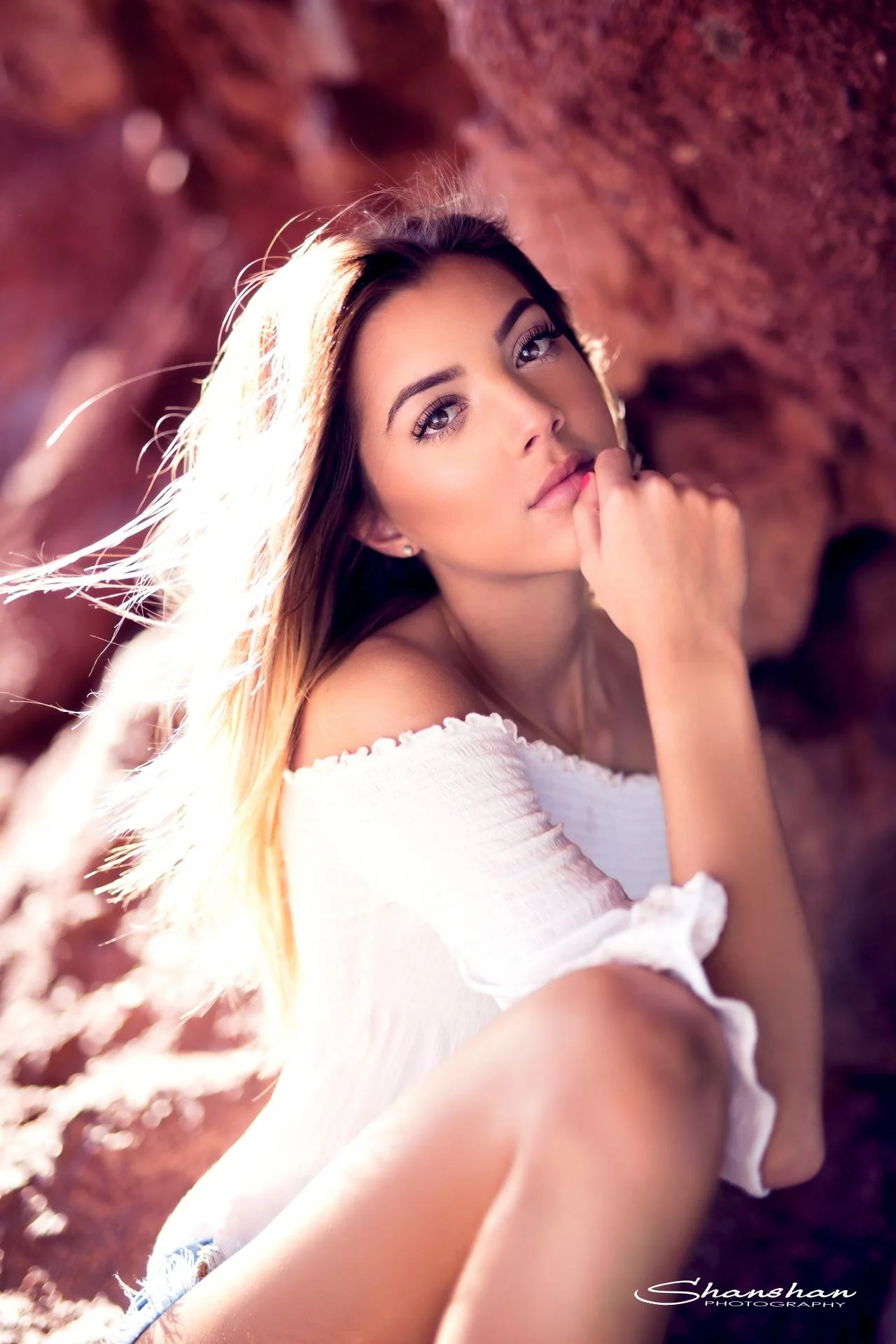 Close-up of a young woman with long hair, lying outdoors on rocks, with sunlight illuminating her face, wearing a white off-shoulder top.