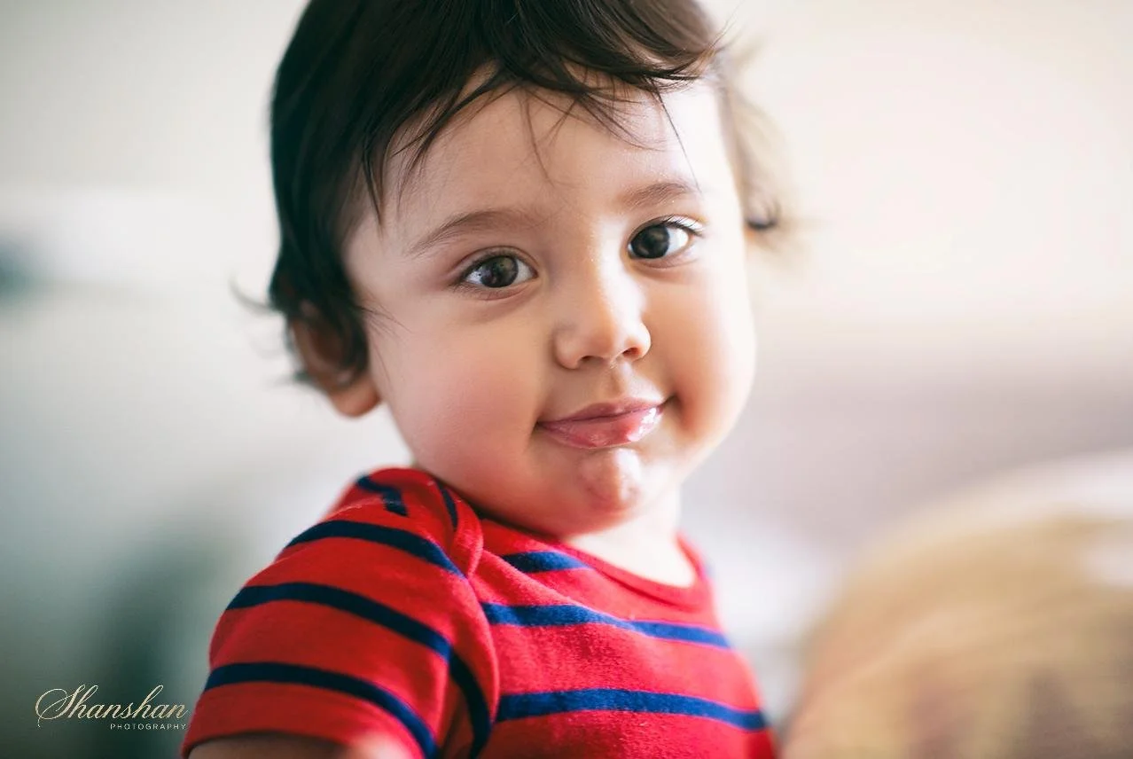 A young child with dark hair, big eyes, and a slight smile, wearing a red and blue striped shirt, looking at the camera.