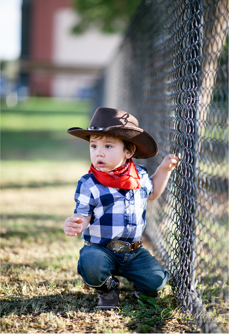 Young child dressed as a cowboy, wearing a cowboy hat, red bandana, checkered shirt, and jeans, crouching and holding onto a chain-link fence outdoor.