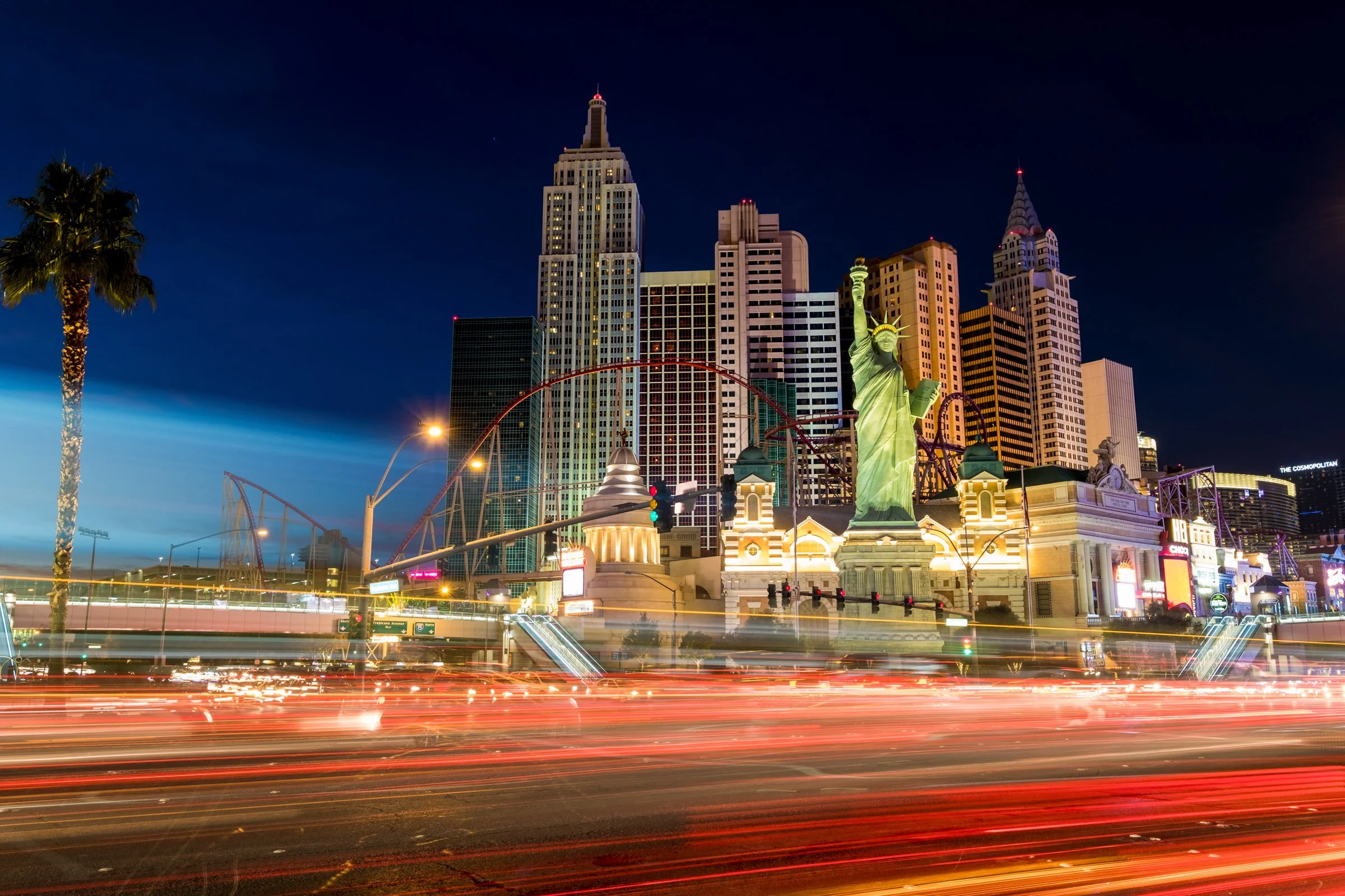 Nighttime view of the Las Vegas Strip with various buildings, including replicas of the Empire State Building and the Statue of Liberty, with light trails from moving vehicles in the foreground.