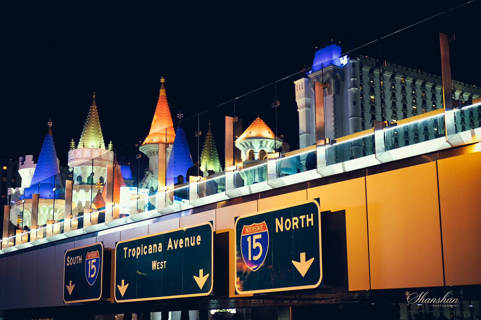 Nighttime city street scene with colorful castle-like structures atop a building, visible above highway signs for Interstate 15 North, Tropicana Avenue, and Las Vegas Boulevard South.