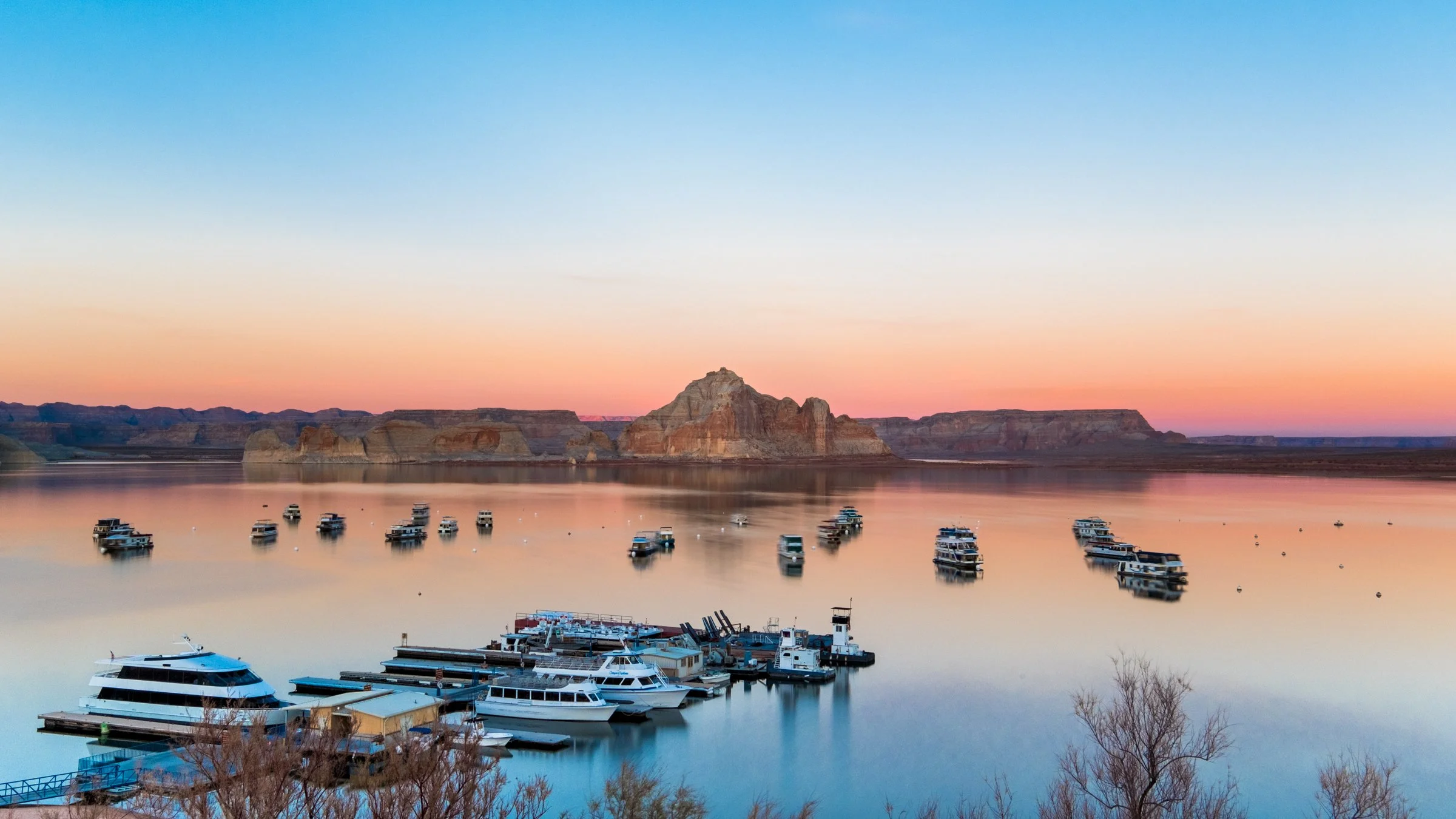 View of Lake Powell at sunset with boats docked at the marina and red rock formations in the background.