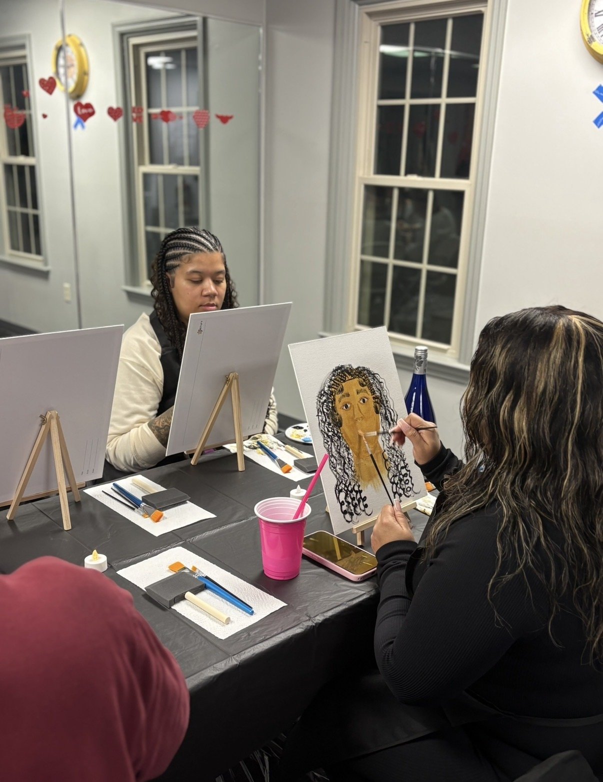 People painting portraits at an indoor art class, with a woman focused on her canvas and another woman painting a woman with curly hair on a smaller canvas.