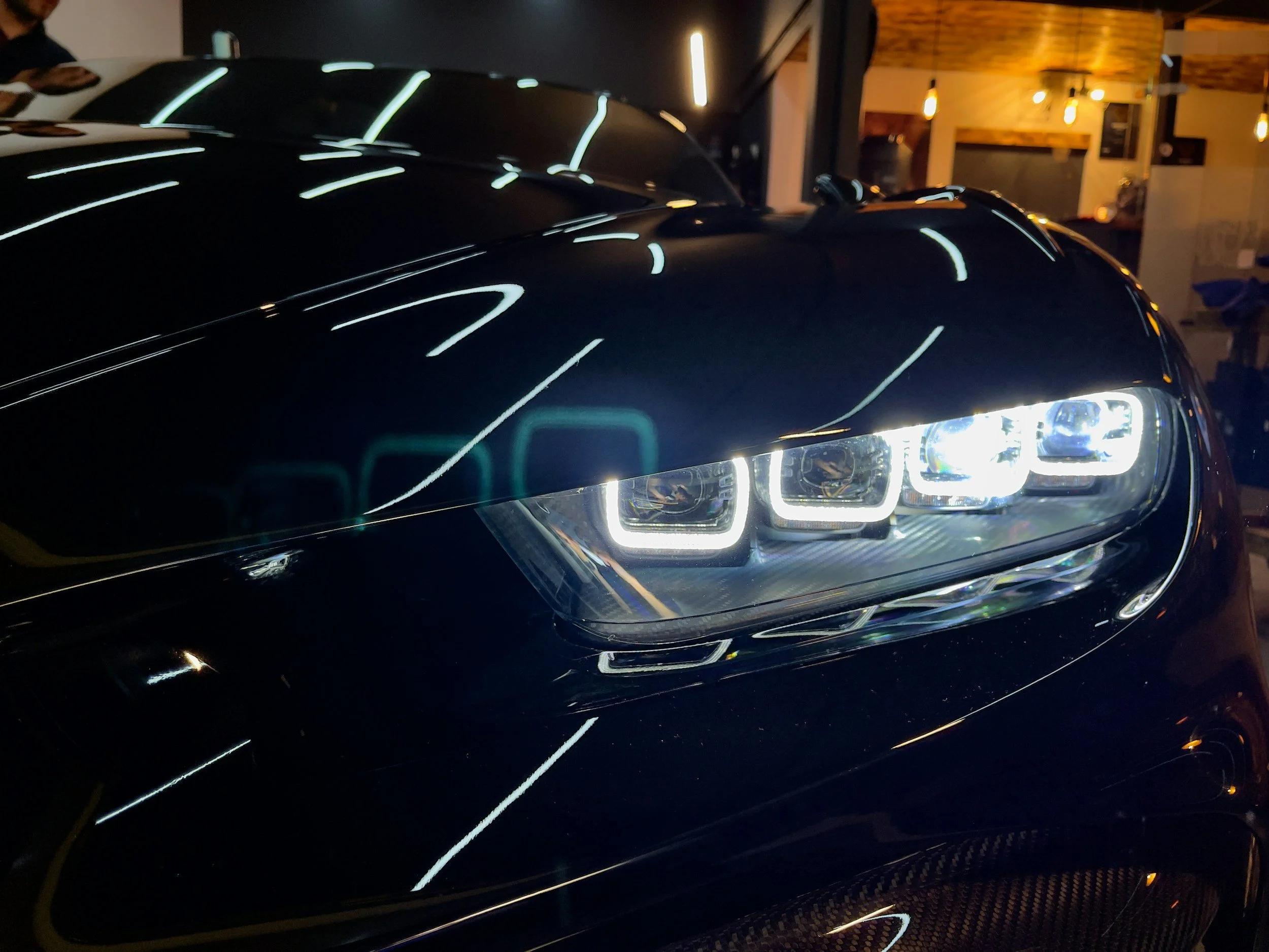 Close-up of a black luxury car with illuminated headlights in a dimly lit indoor space.