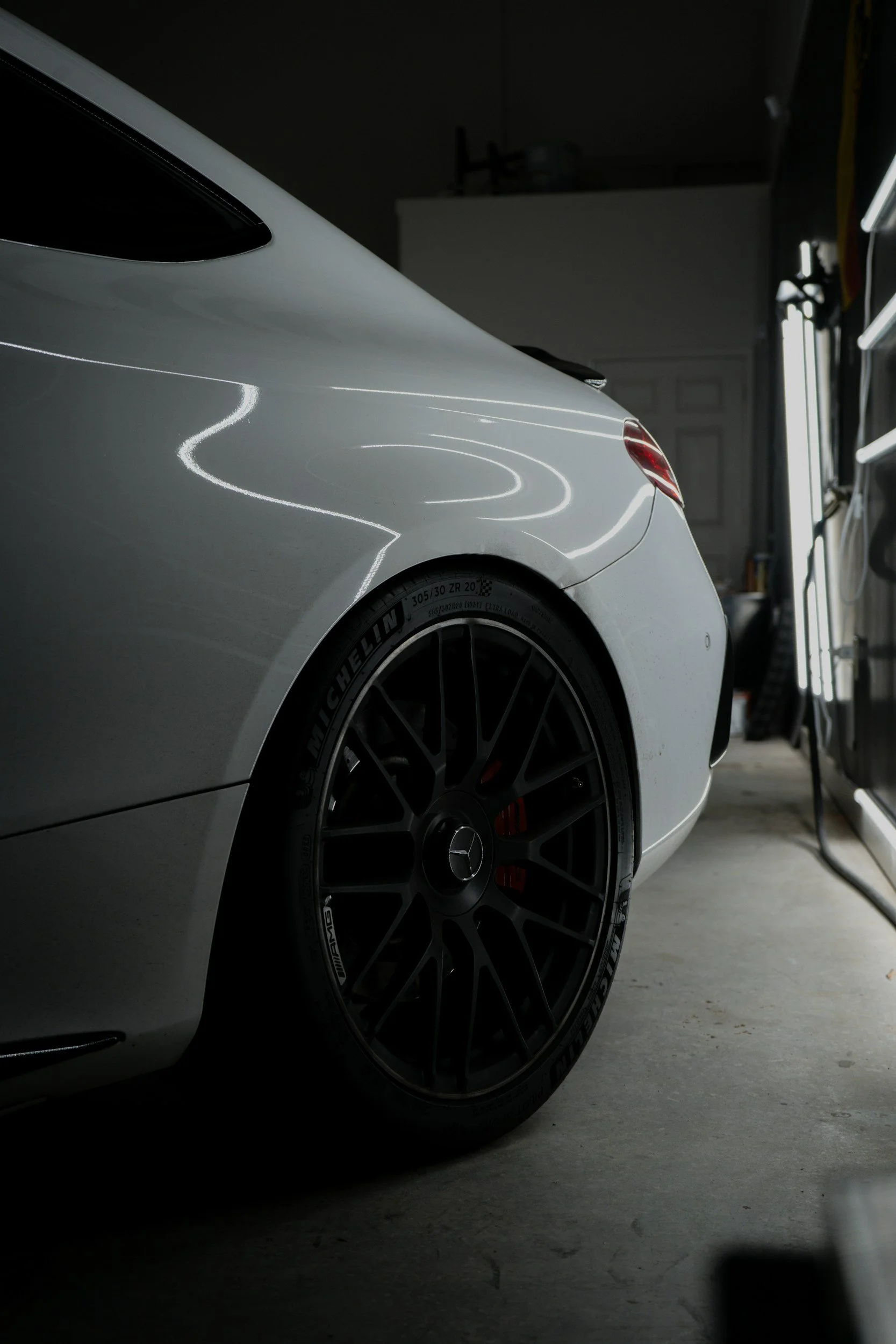 Close-up of a white luxury sports car in a garage with a black tire and silver alloy wheel, reflected under bright indoor lighting.