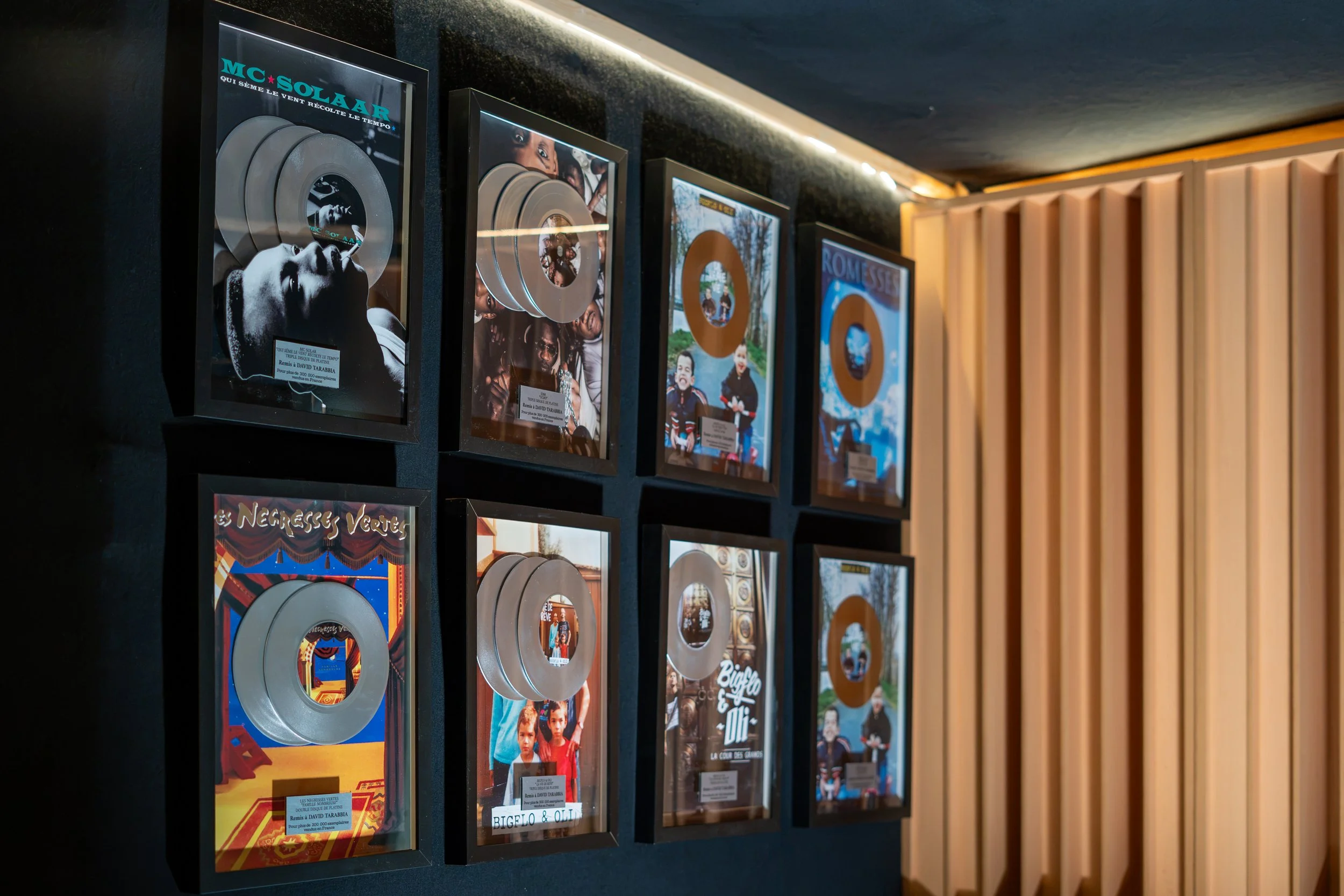 Cadre avec plusieurs affiches de prix de disques en or et en argent, certains présentant des photos de personnes ou d'illustrations, disposées sur un mur noir avec des bandes de bois clair à droite, dans une salle d'exposition ou une boutique musicale.